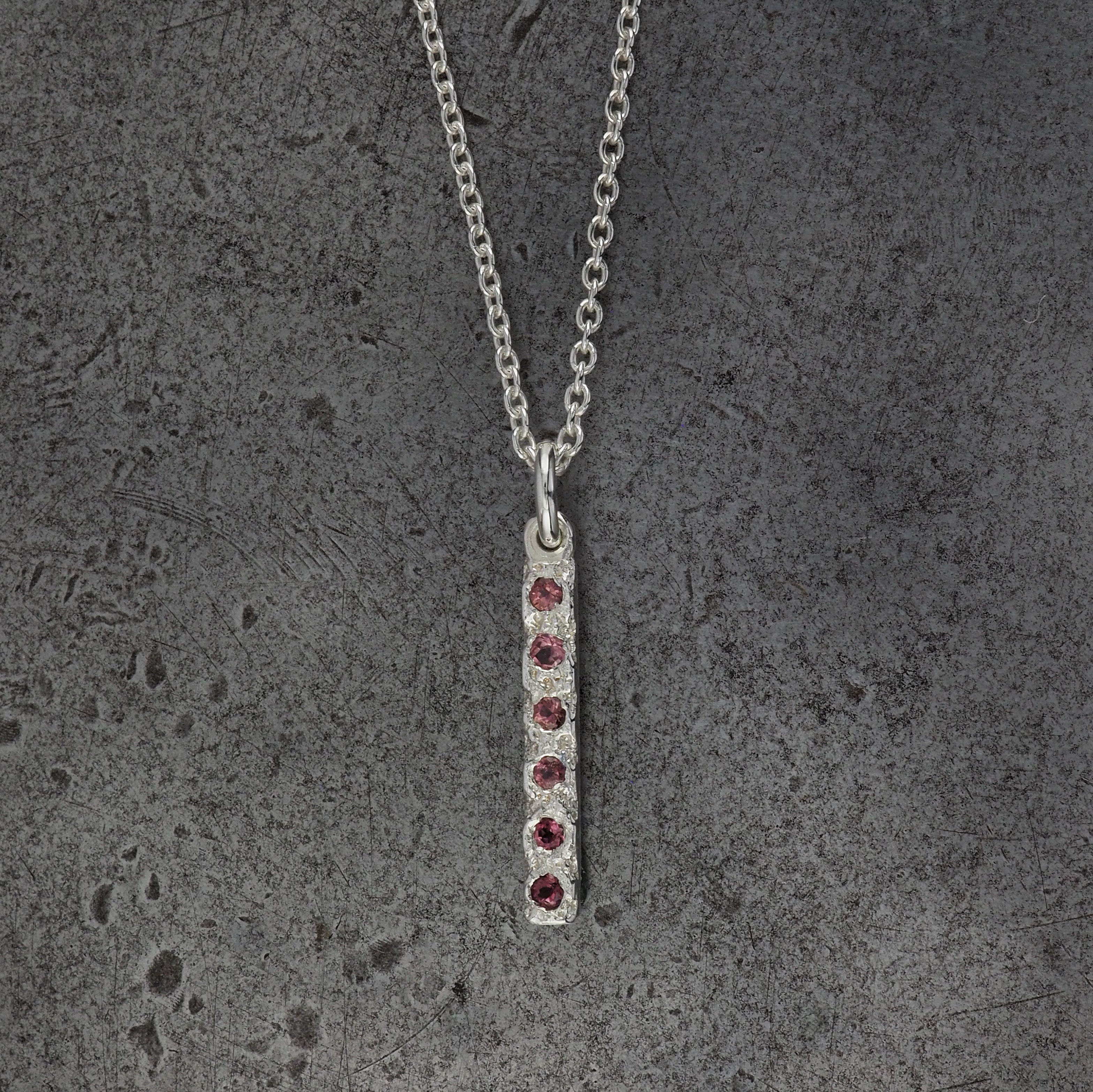 A Sterling silver chain necklace with a small textured rectangle bar pendant with 6 tourmaline birthstones on a steel textured background.