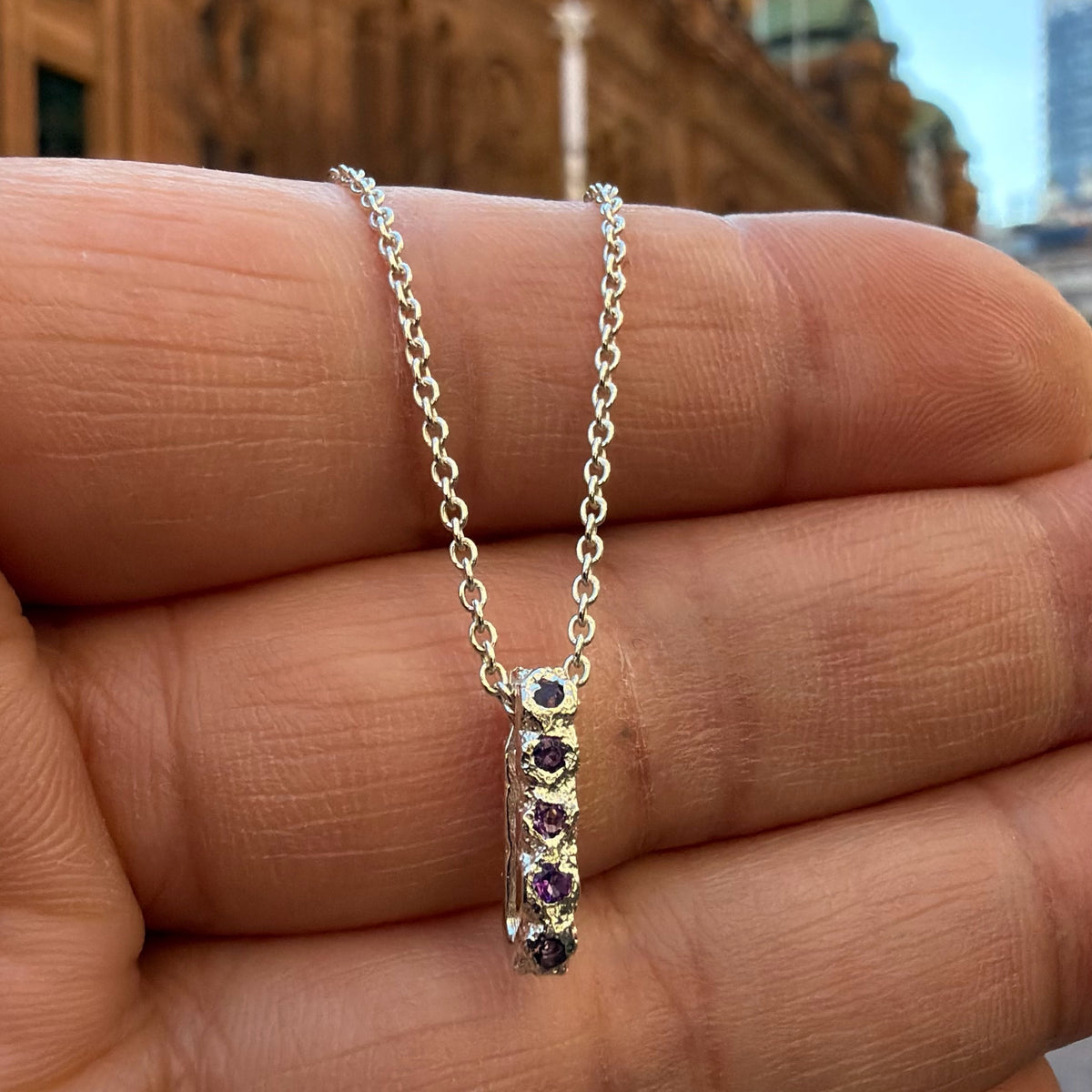 A Sterling silver chain necklace with a small textured oval bead pendant with 12 amethyst birthstones, 5 on the front going all they way around, on the hand for scale