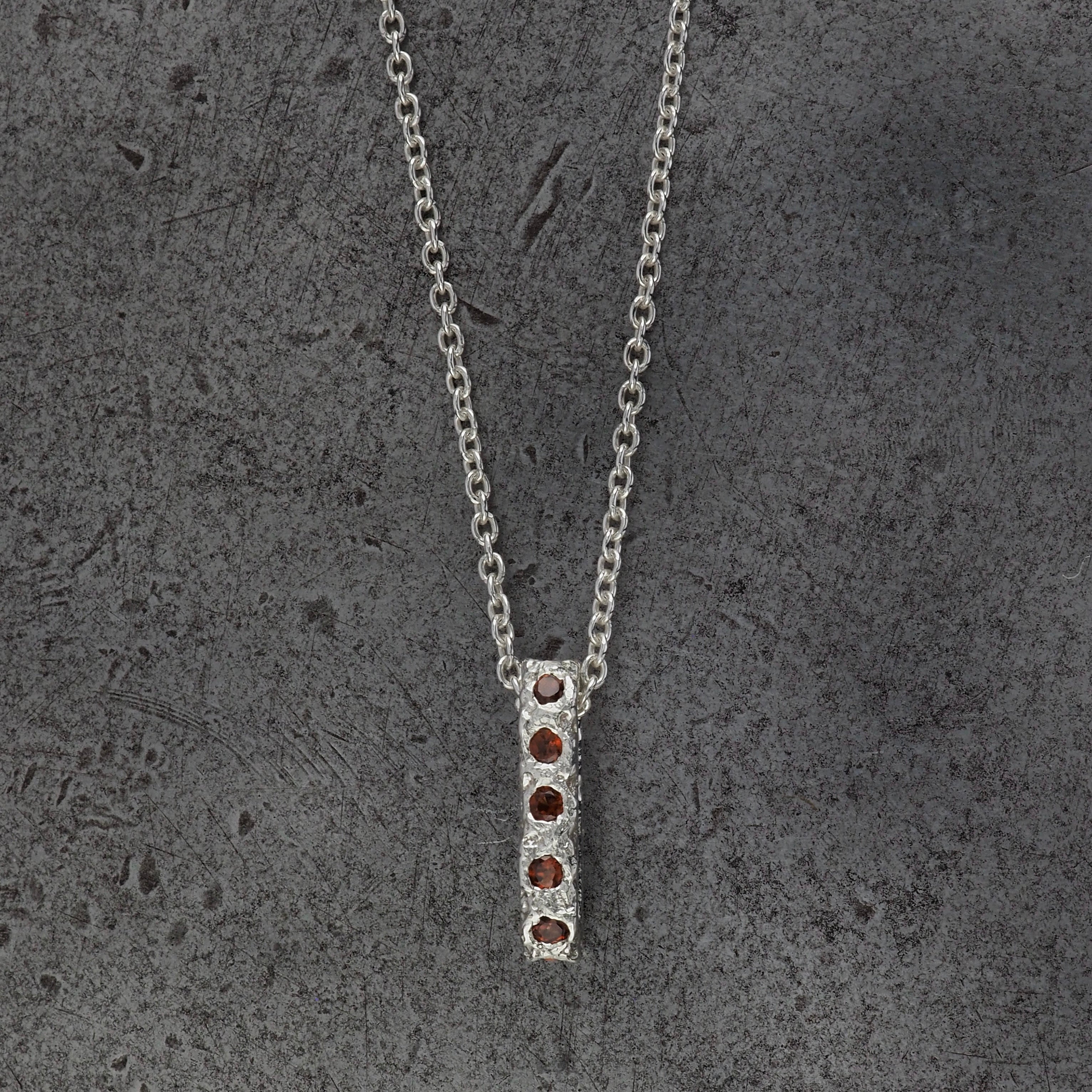 A Sterling silver chain necklace with a small textured oval bead pendant with garnet birthstones, displayed on a steel textured background.