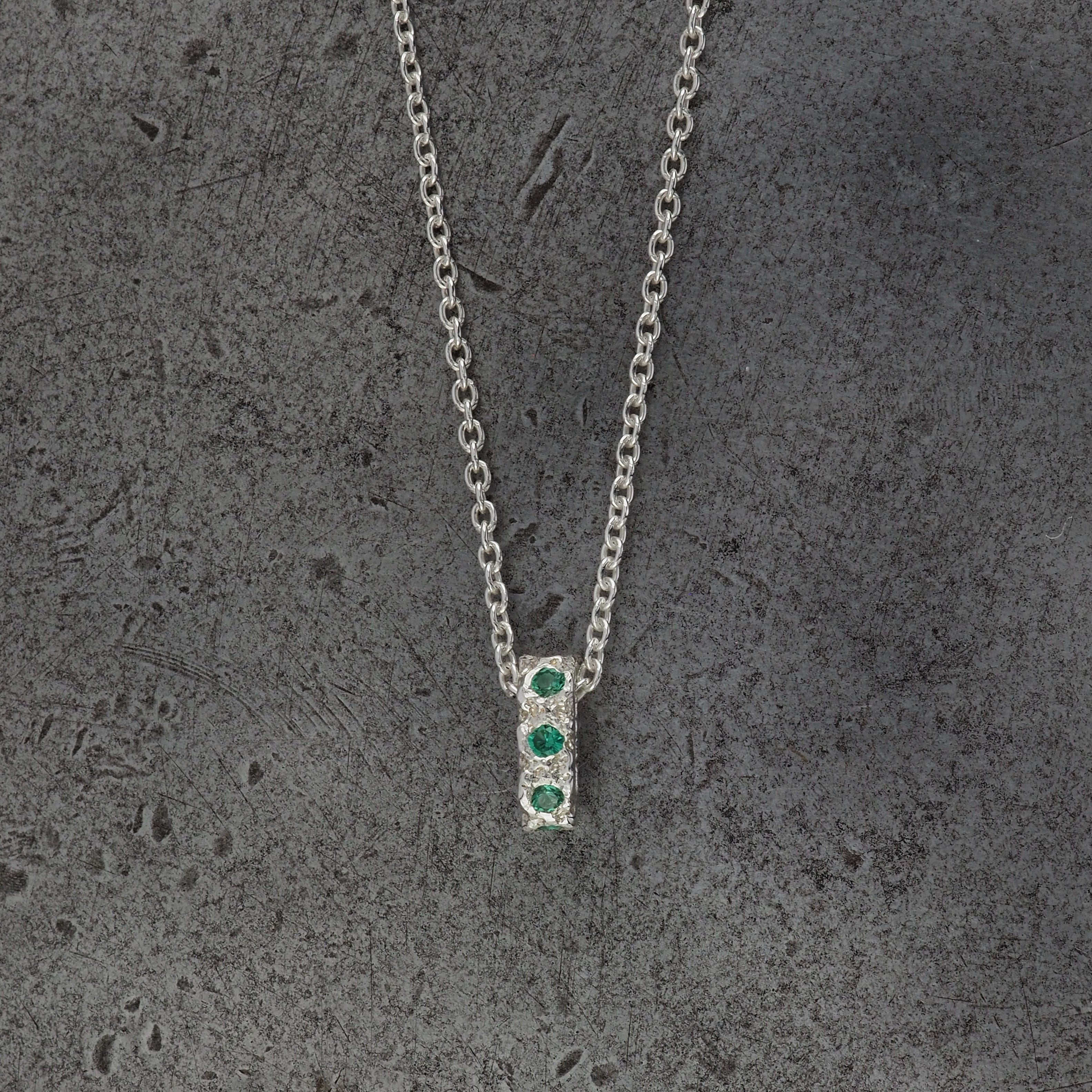 A Sterling silver chain necklace with a small textured oval bead pendant with 3 emerald birthstones on the front going all the way around, displayed on a steel textured background.