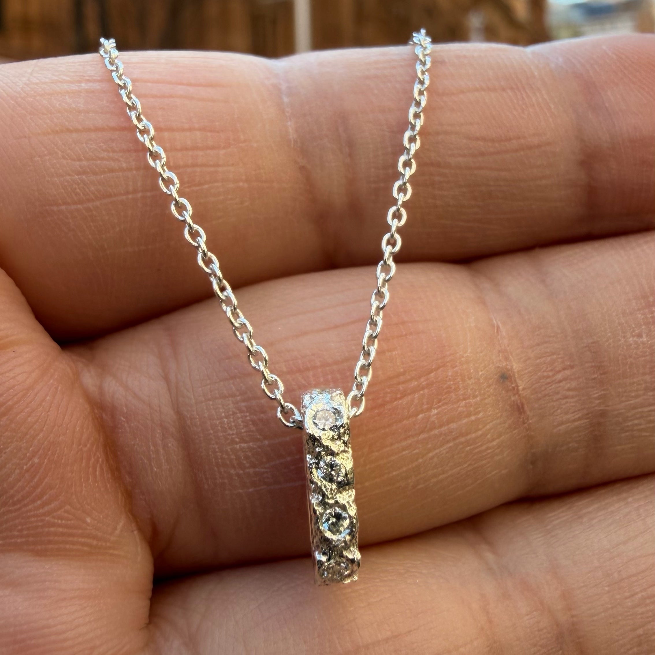 A Sterling silver chain necklace with a small textured oval bead pendant with 10 diamond birthstones, 4 on the front going all they way around, on the hand for scale