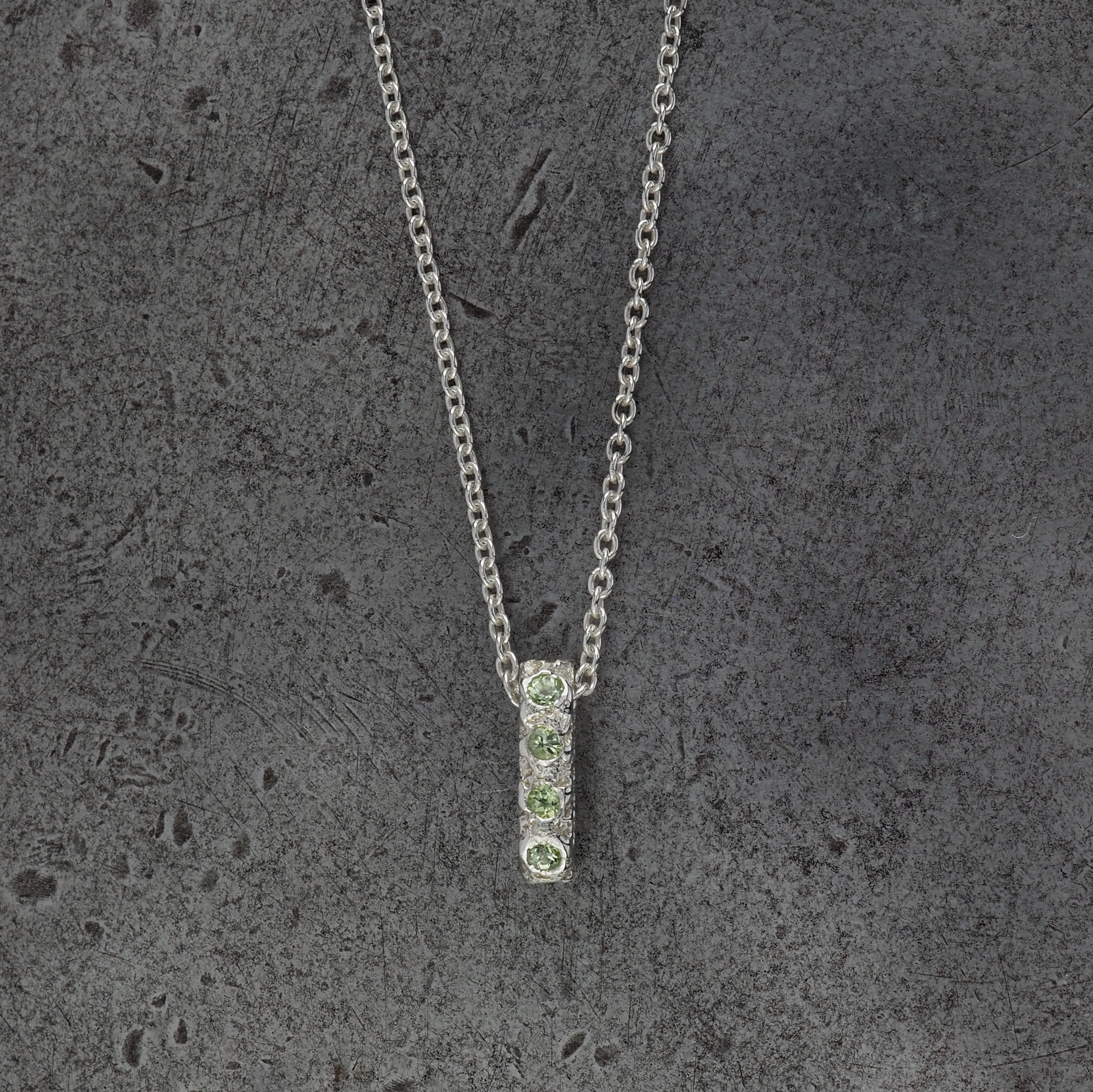 A Sterling silver chain necklace with a small textured oval bead pendant with 10 peridot birthstones, displayed on a steel textured background.