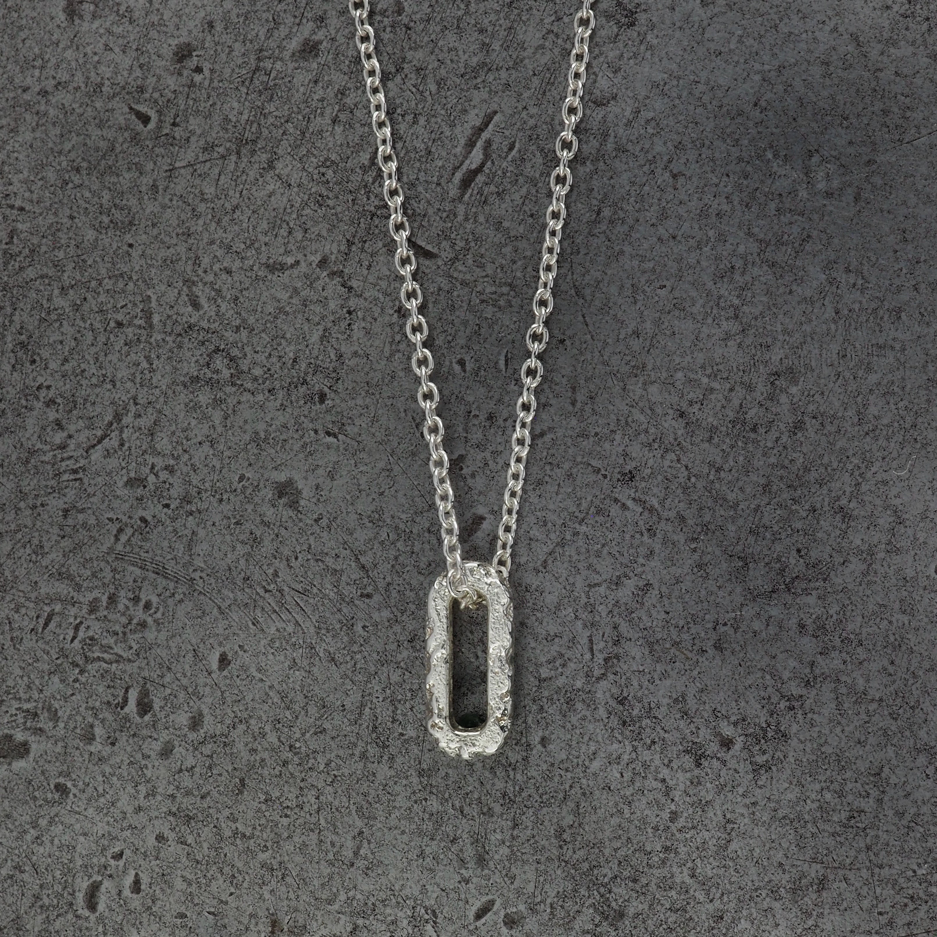 A Sterling silver chain necklace with a small textured oval bead pendant side profile, displayed on a steel background.