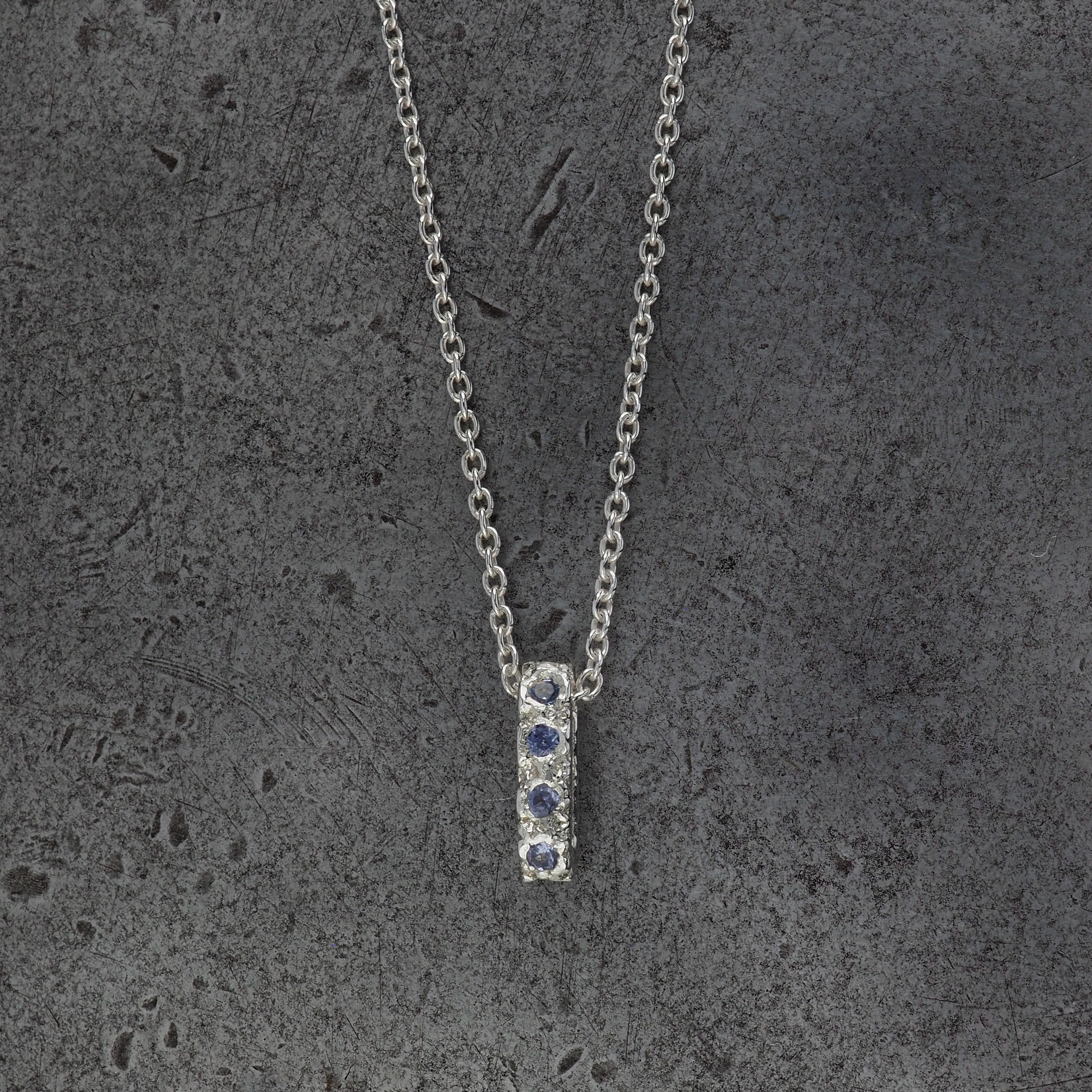 A Sterling silver chain necklace with a small textured oval bead pendant with 10 tanzanite birthstones, displayed on a steel textured background.