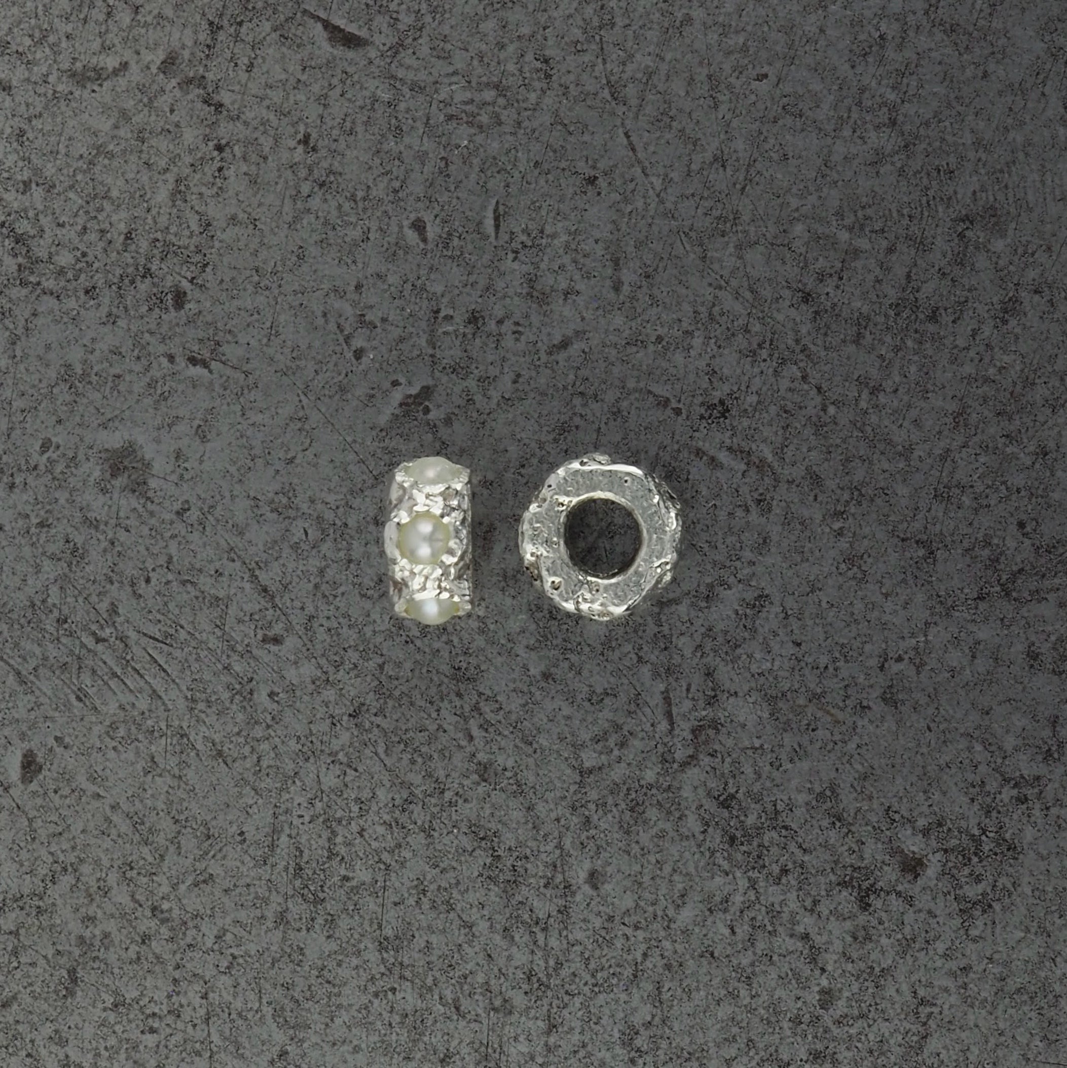 Two small textured cylindrical bead pendants, one pearl and one with aquamarine birthstones side profile, displayed on a steel background.