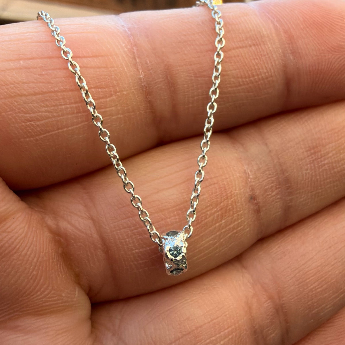 A Sterling silver chain necklace with a small textured cylindrical bead pendant with 6 aquamarine birthstones going all the way around, on the hand for scale