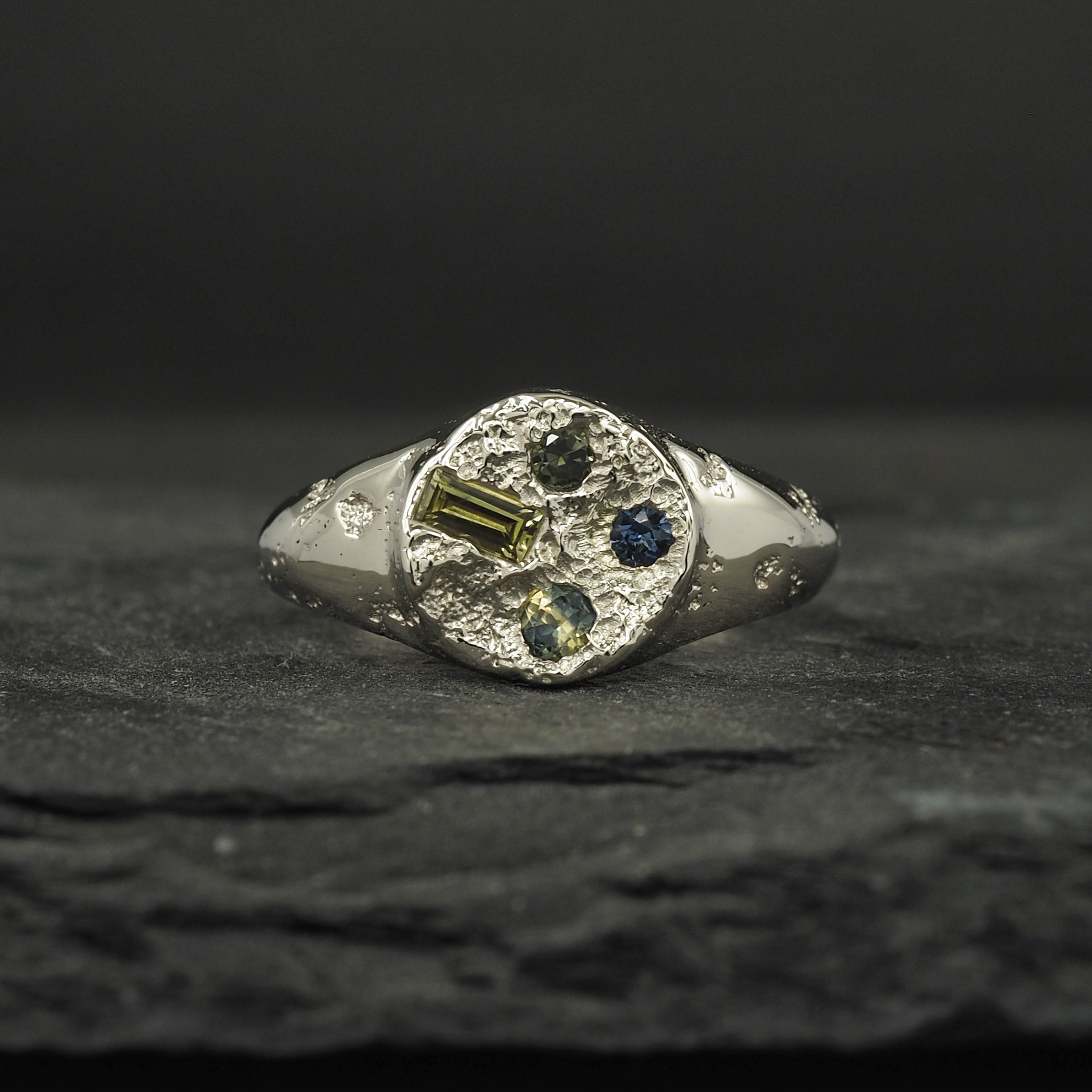 Small textured round signet ring in 9k white gold with a terrain texture that signifies the land from high able, with four cast in place and set Australian sapphires