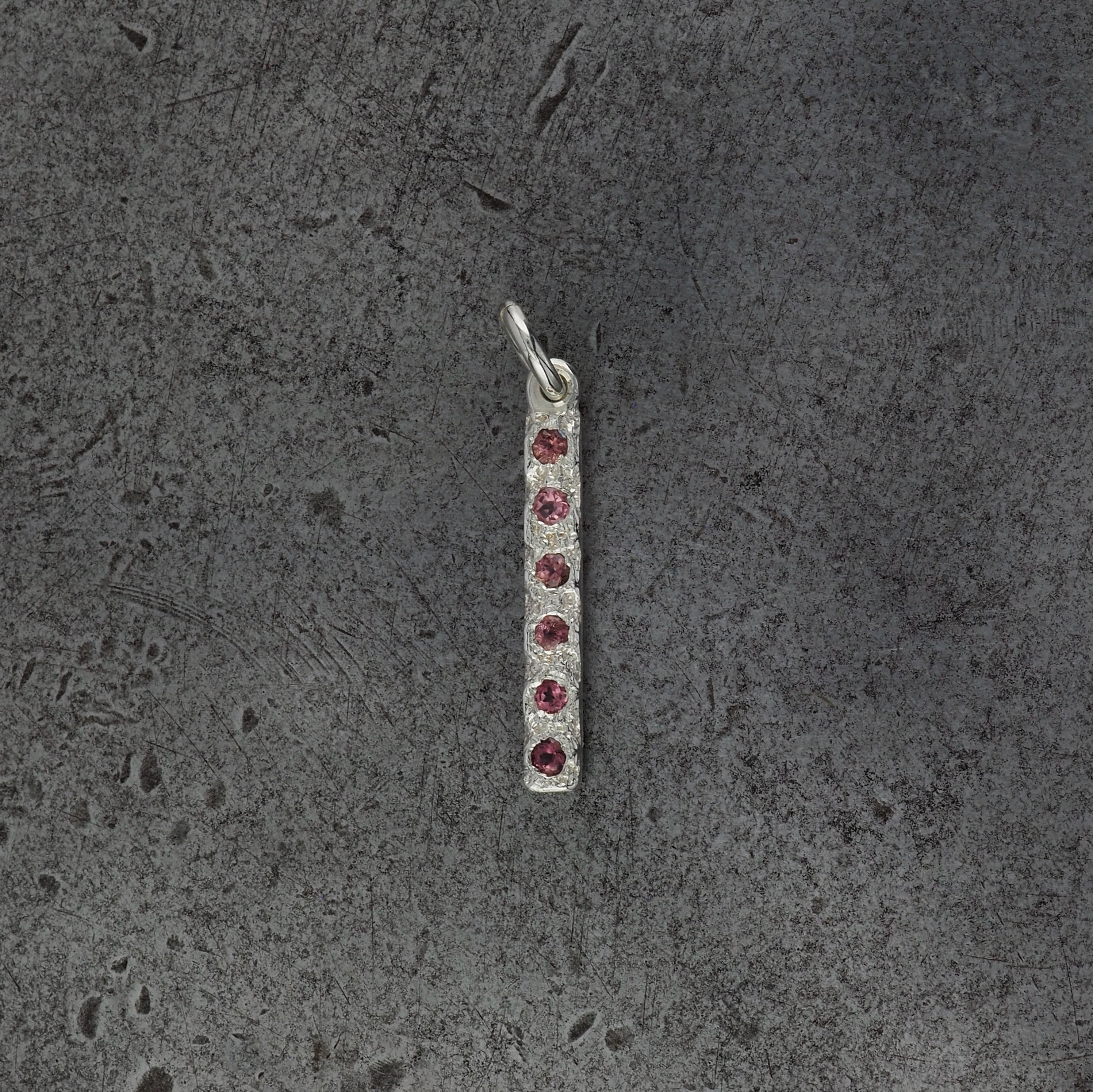 A small textured rectangle bar pendant with 6 tourmaline birthstones on a steel textured background.
