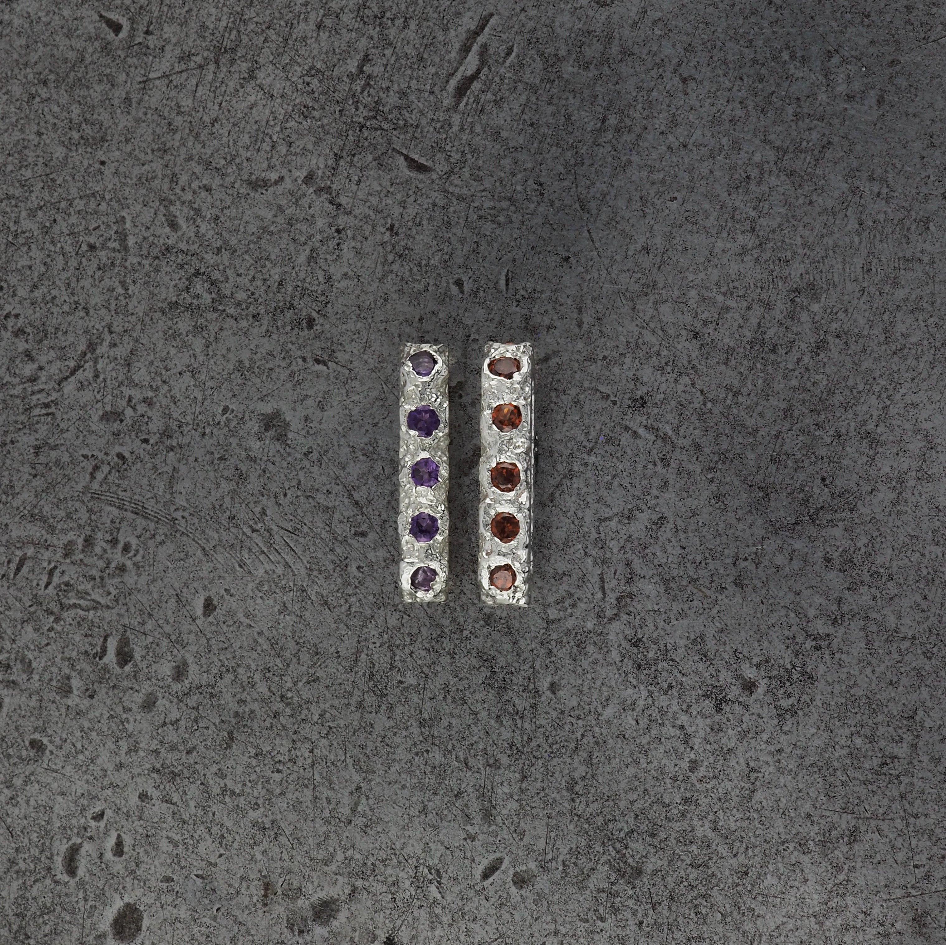 Two small textured oval bead pendants, one with amethyst birthstones and the other with garnet birthstones on a steel textured background.