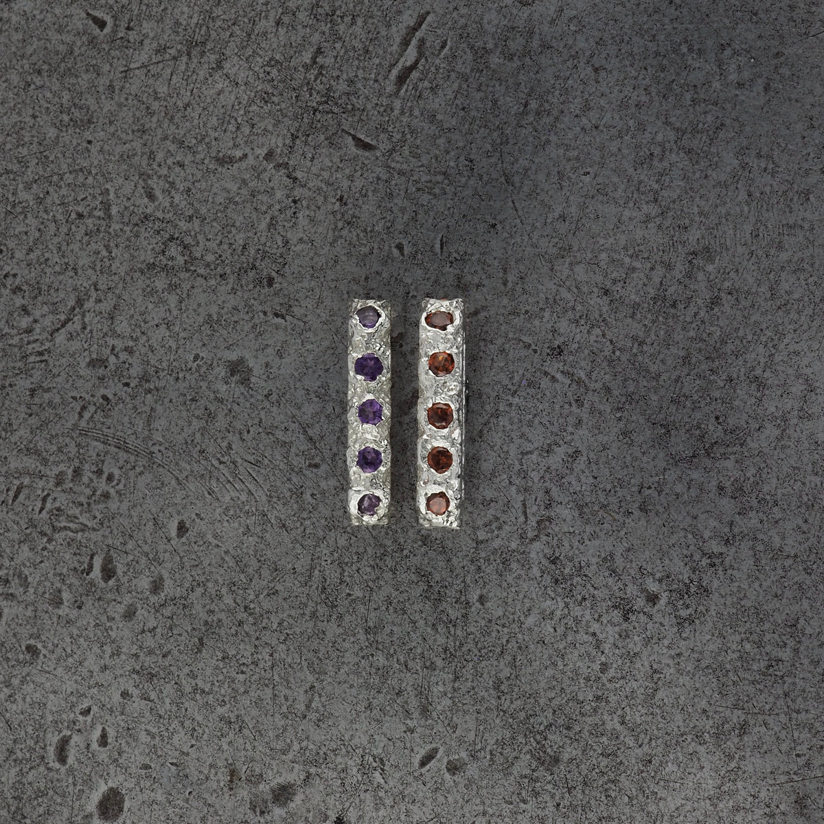 Two small textured oval bead pendants, one with amethyst birthstones and the other with garnet birthstones on a steel textured background.