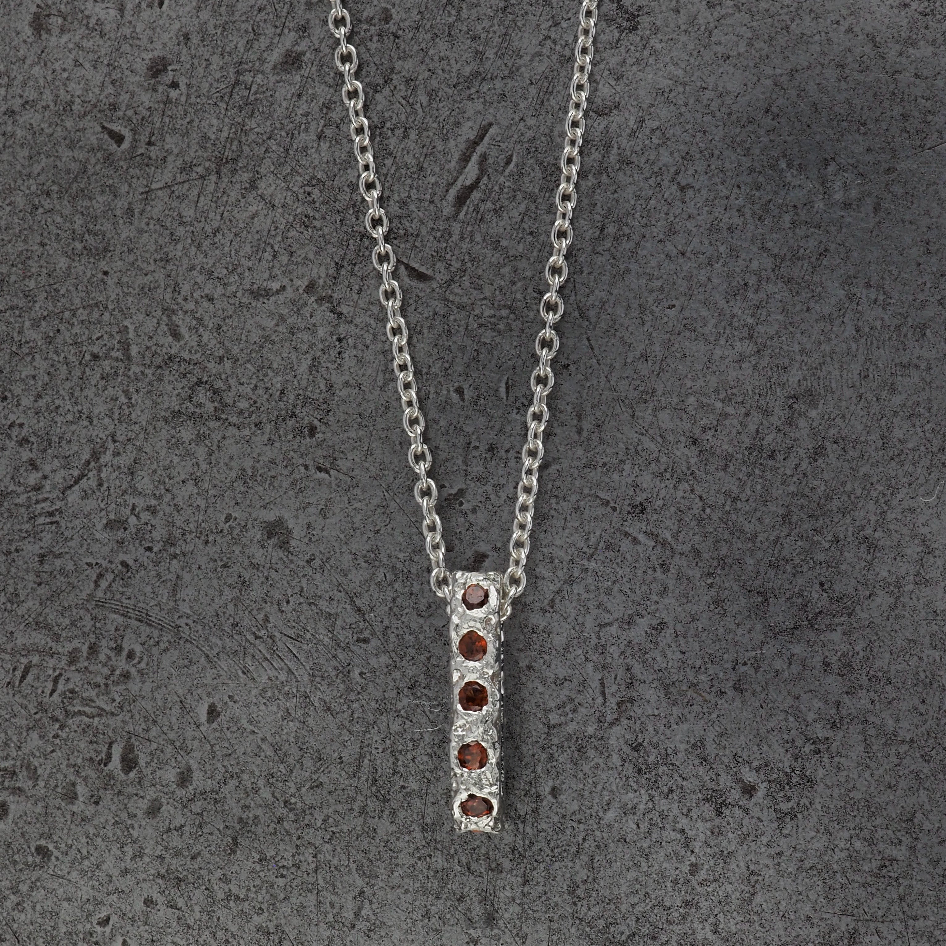 A Sterling silver chain necklace with a small textured oval bead pendant with garnet birthstones, displayed on a steel textured background.