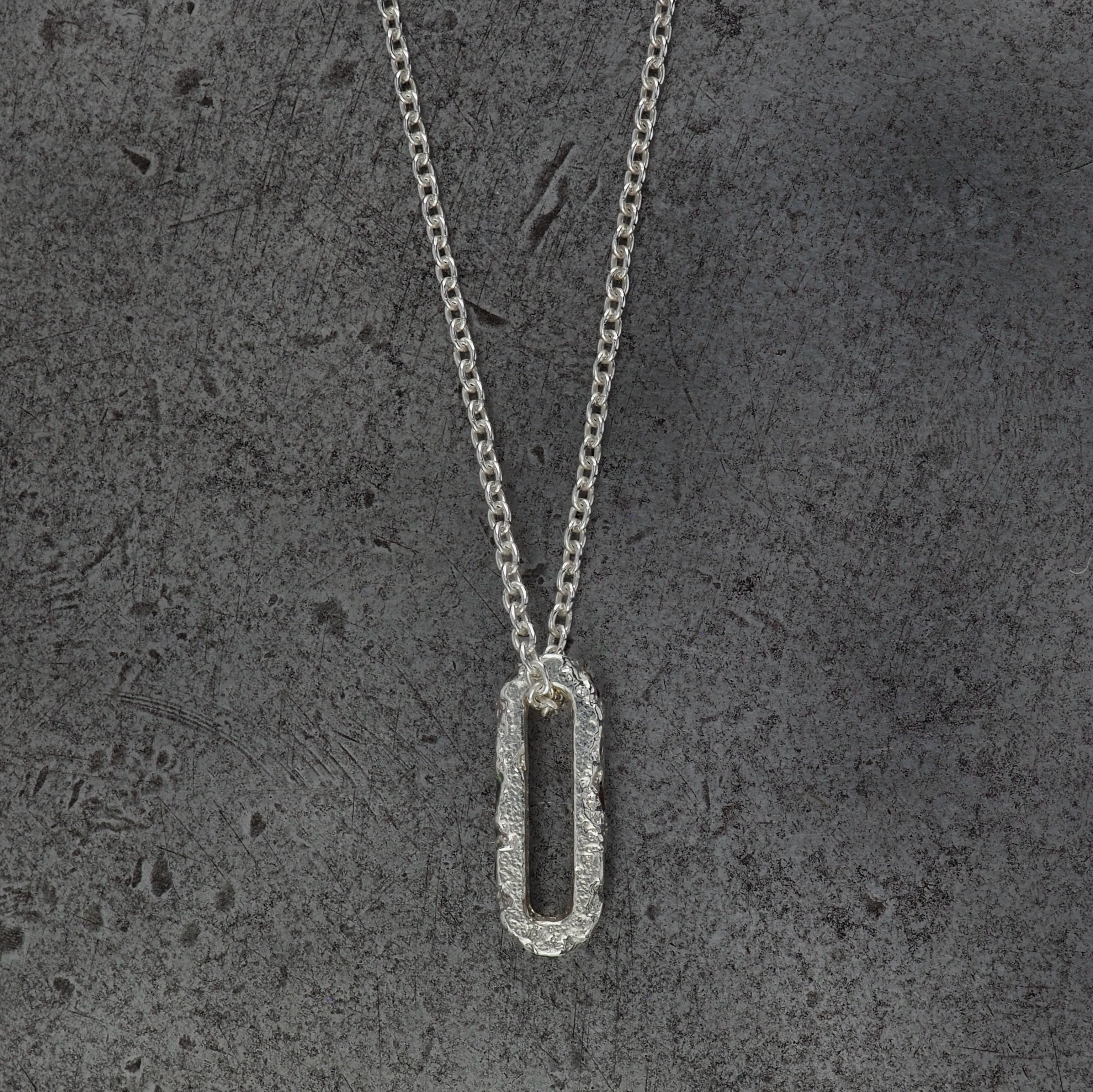 A Sterling silver chain necklace with a small textured oval bead pendant side profile, displayed on a steel textured background.
