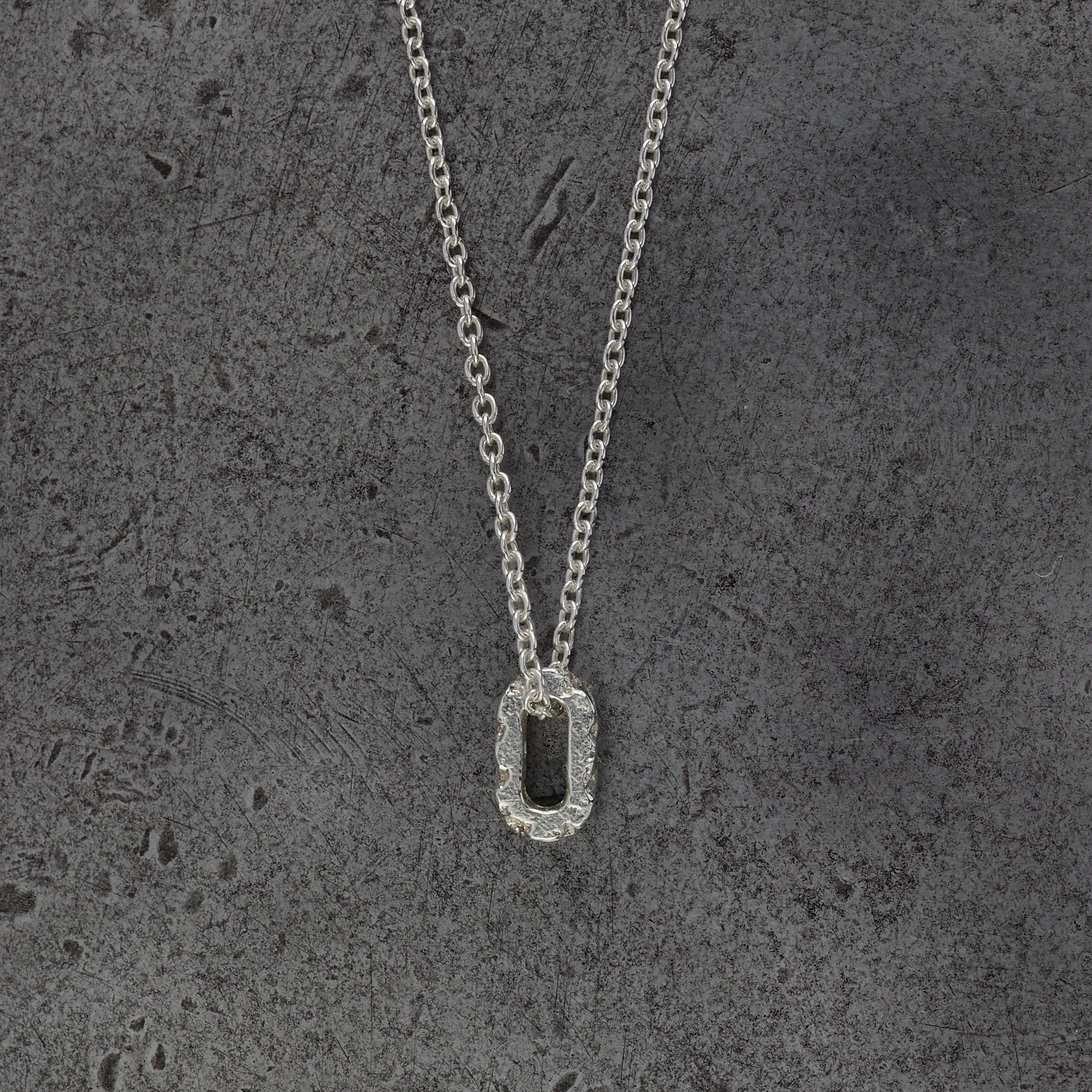 A Sterling silver chain necklace with a small textured oval bead pendant side profile, displayed on a steel textured background.