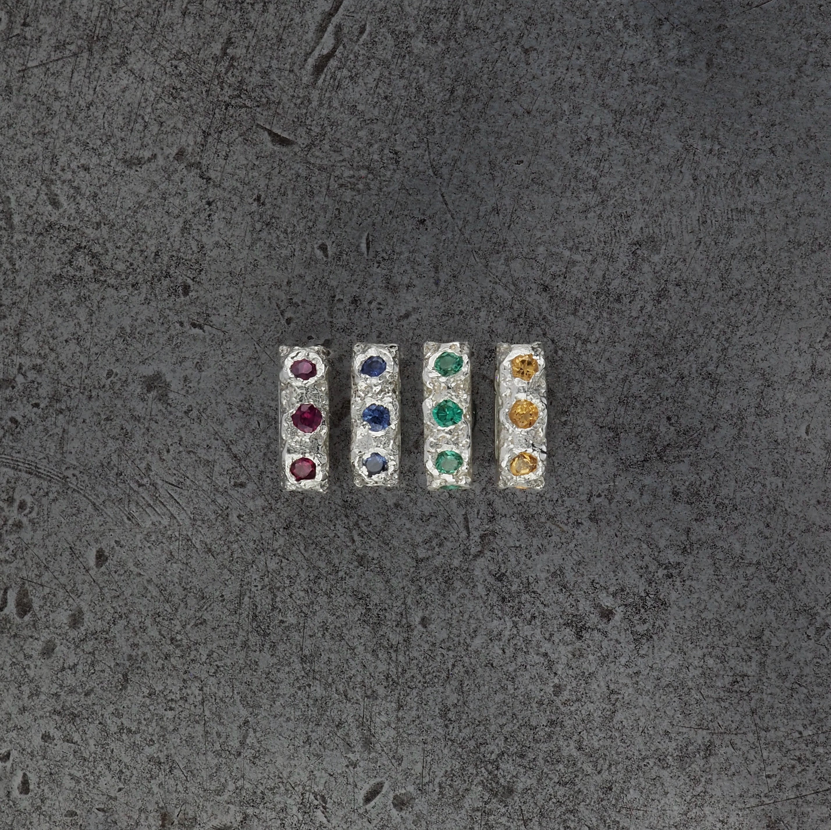 Four small textured oval bead pendants, one with ruby, sapphire, emerald, citrine birthstones on a steel textured background.