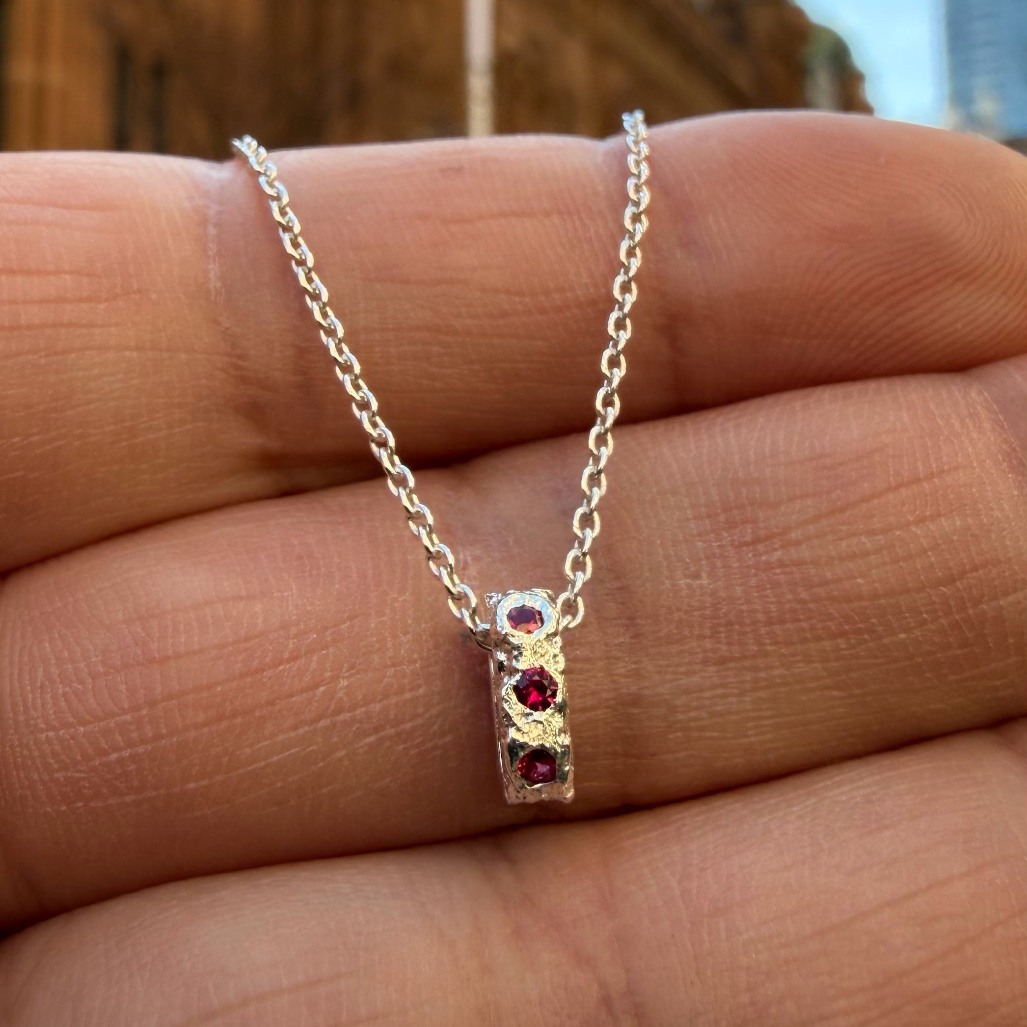 A Sterling silver chain necklace with a small textured oval bead pendant with 8 ruby birthstones, 3 on the front going all they way around, on the hand for scale