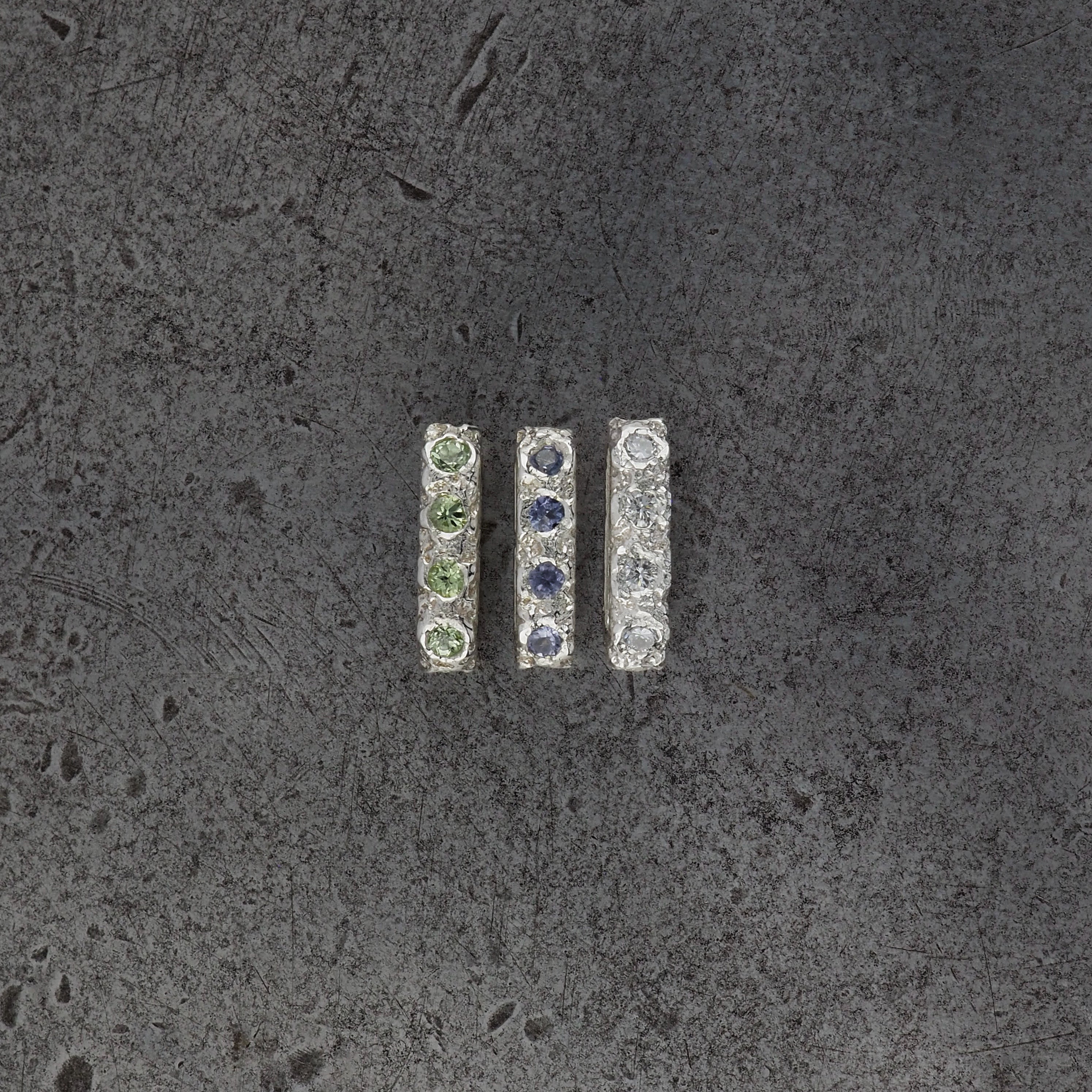 Three small textured oval bead pendants, one with peridot, tanzanite, diamond birthstones on a steel textured background.