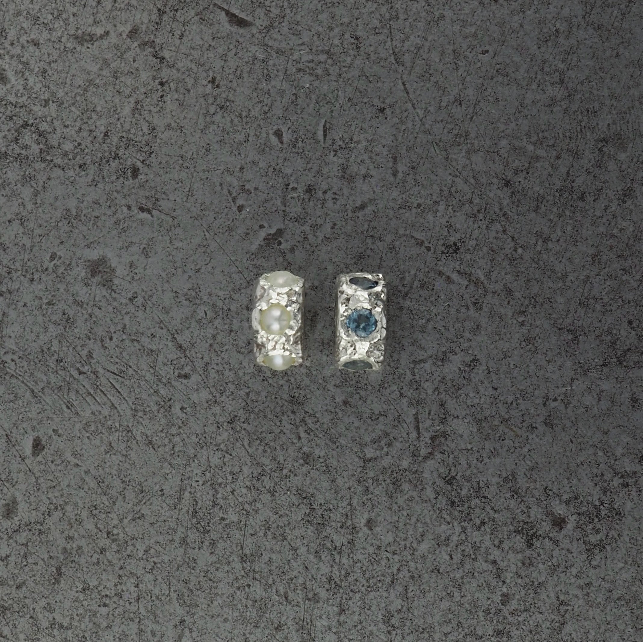 Two small textured cylindrical bead pendants, one with Pearl and one with aquamarine birthstones, displayed on a steel background.