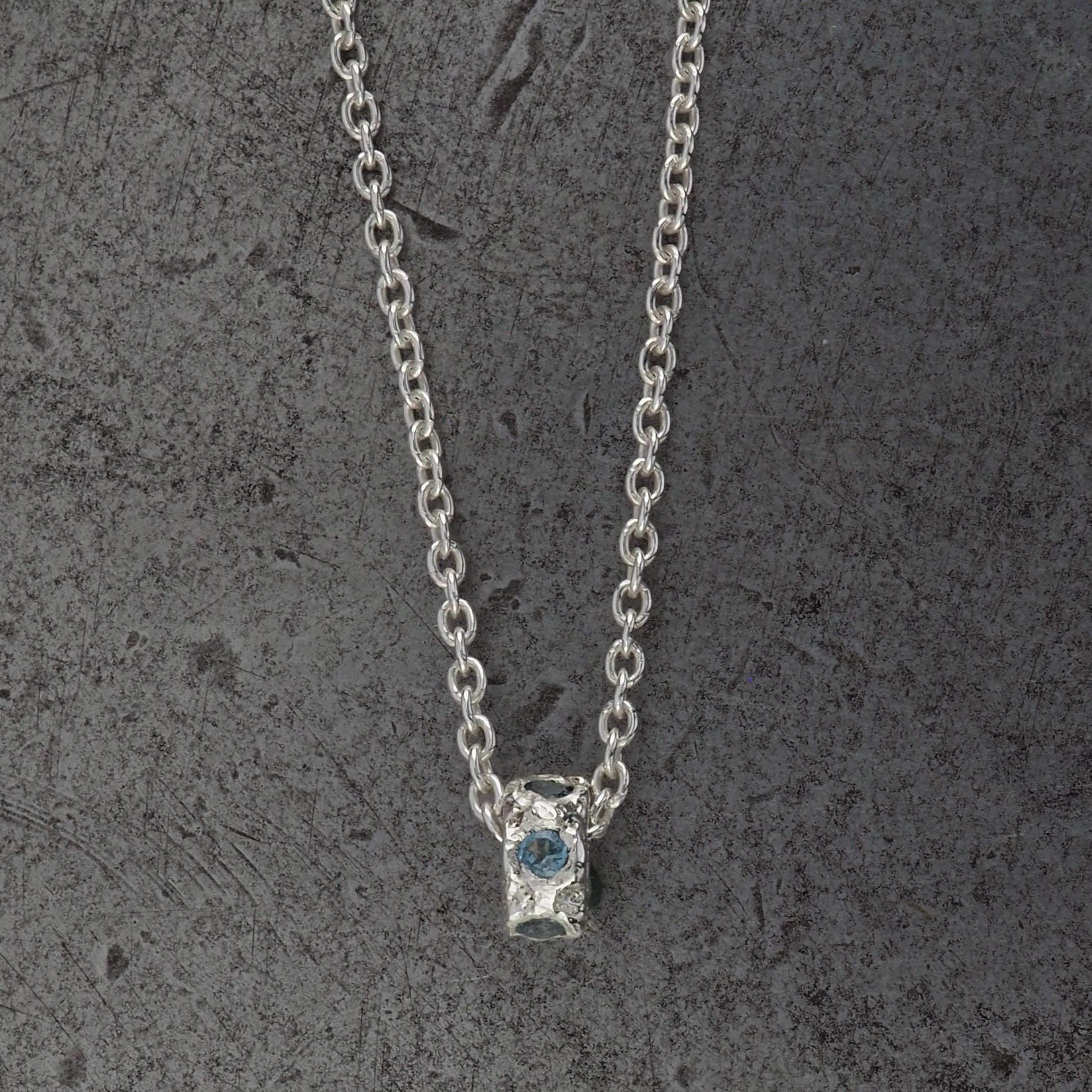 A Sterling silver chain necklace with a small textured cylindrical bead pendant with aquamarine birthstones, displayed on a steel background.