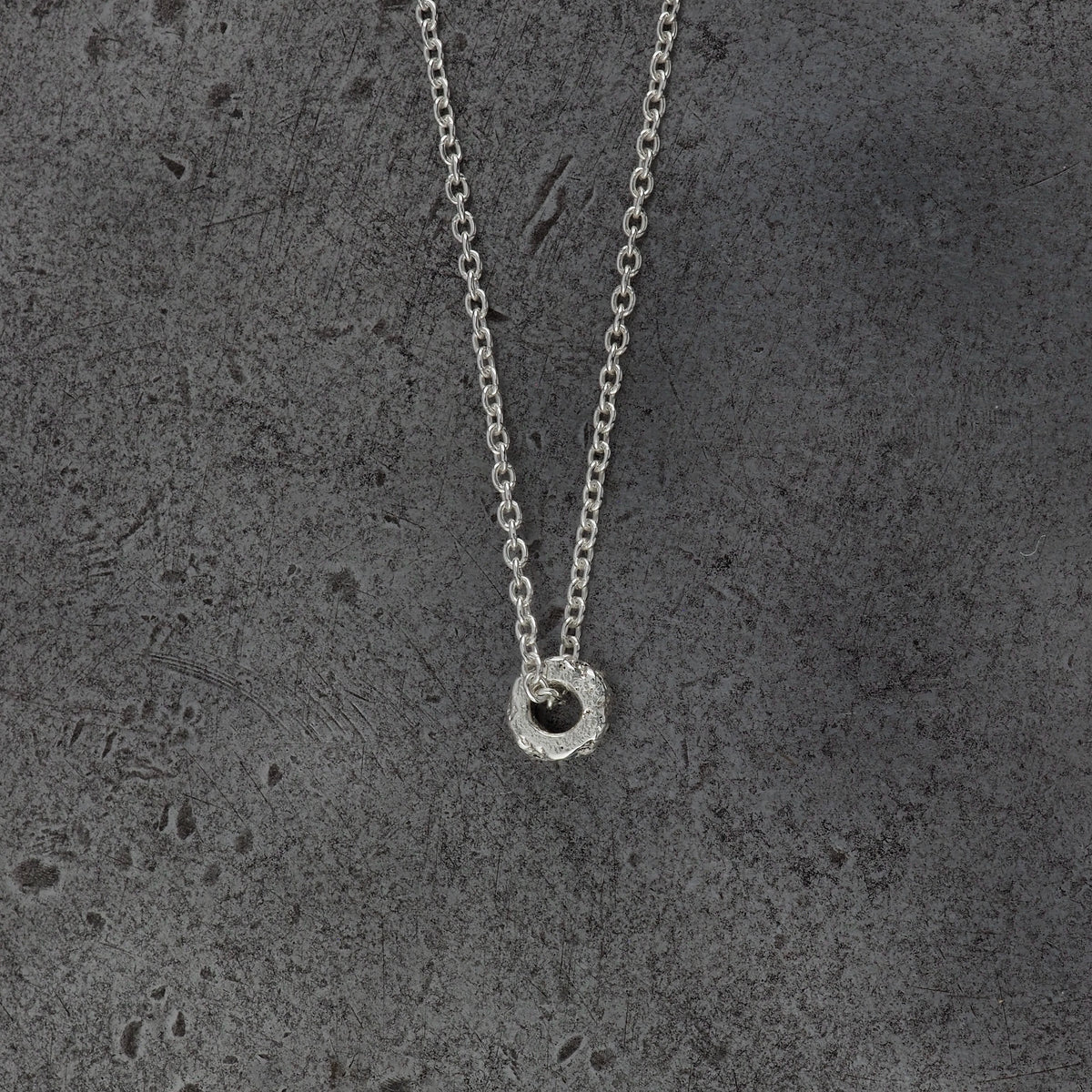 A Sterling silver chain necklace with a small textured cylindrical bead pendant with aquamarine birthstones, side profile displayed on a steel background.