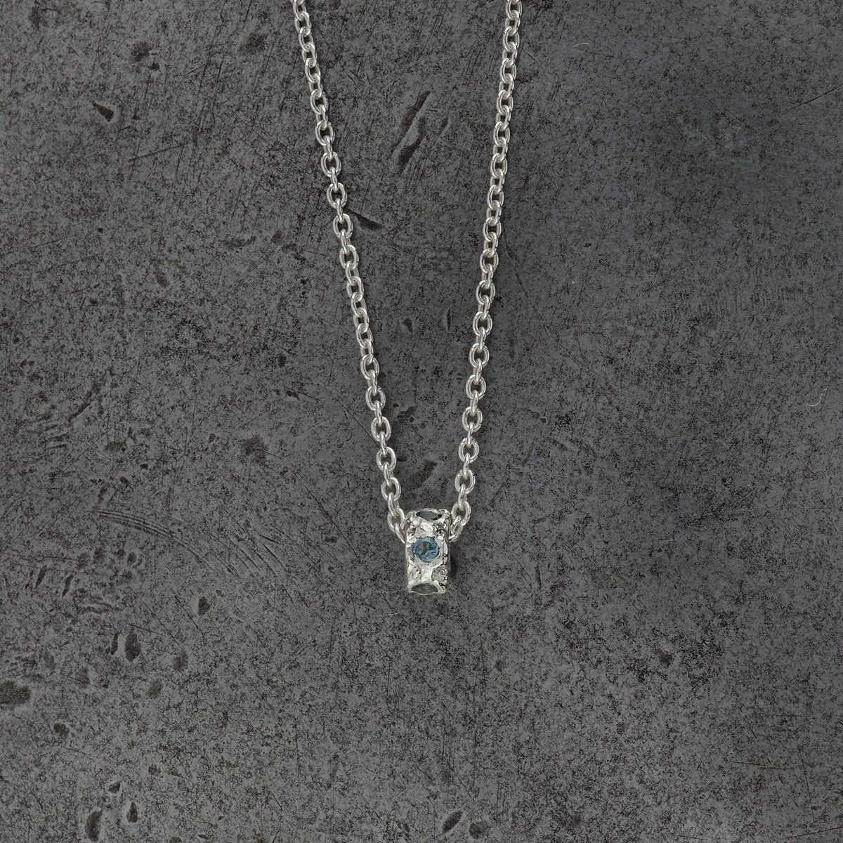 A Sterling silver chain necklace with a small textured cylindrical bead pendant with aquamarine birthstones, displayed on a steel background.