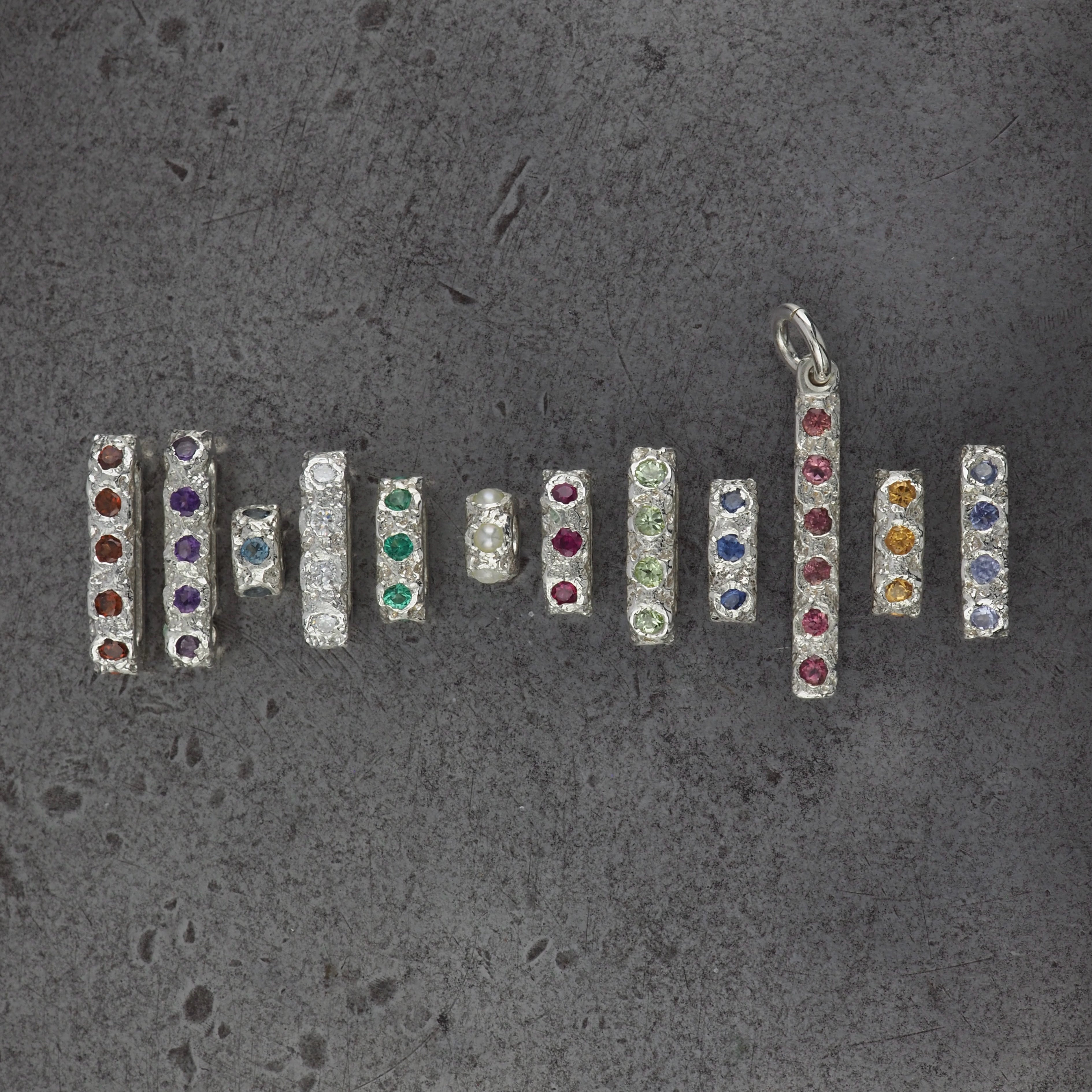 A group of textured pendants in different bead sizes and styles, birthstone pendants going from left to right, January to December. Garnet, amethyst, aquamarine, diamond, emerald, pearl, ruby, peridot, sapphire, tourmaline, citrine and tanzanite. 